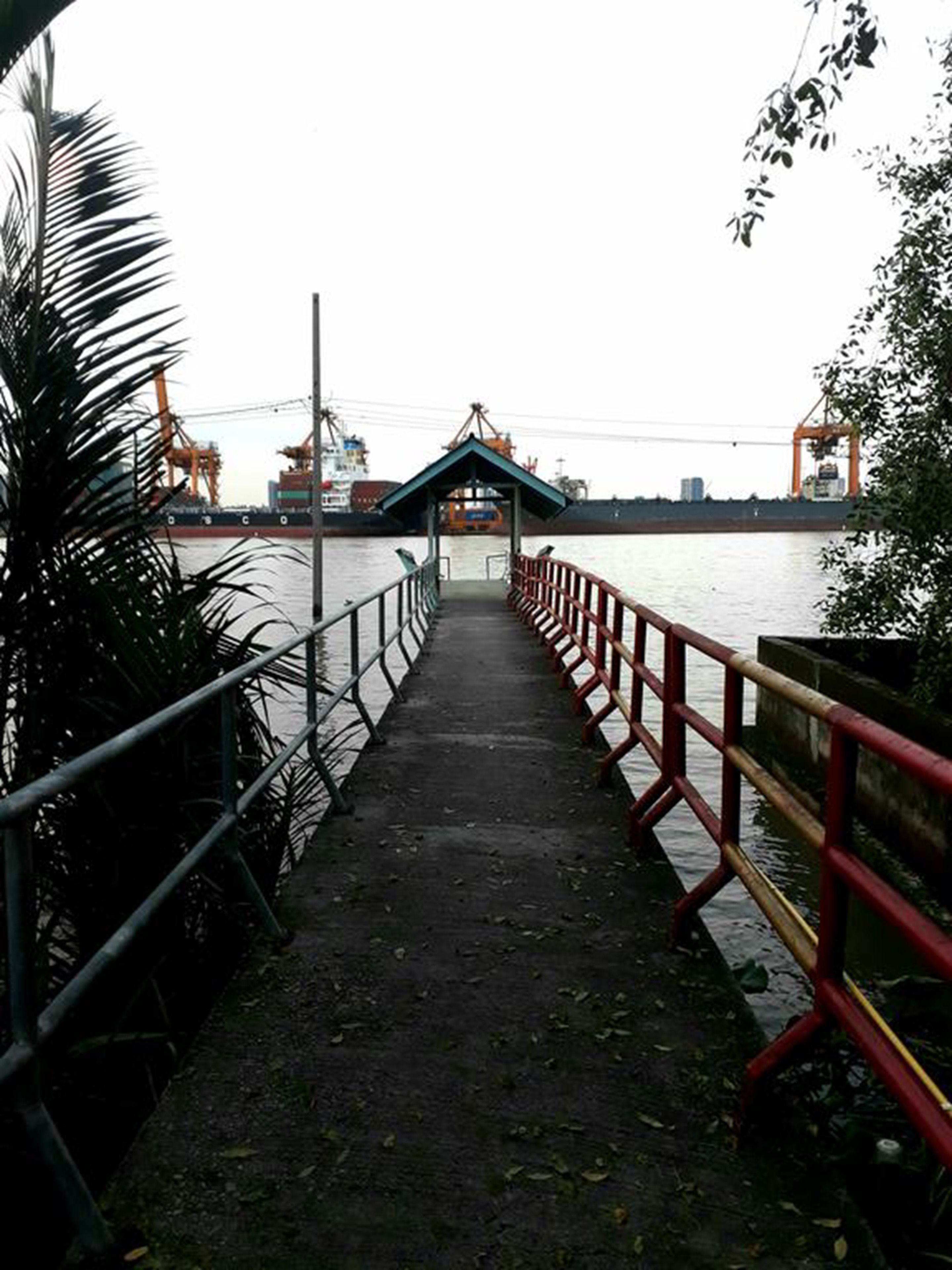 A concrete walkway leading to a covered shelter with a metal railing on both sides, overlooking a body of water with industrial structures in the background.