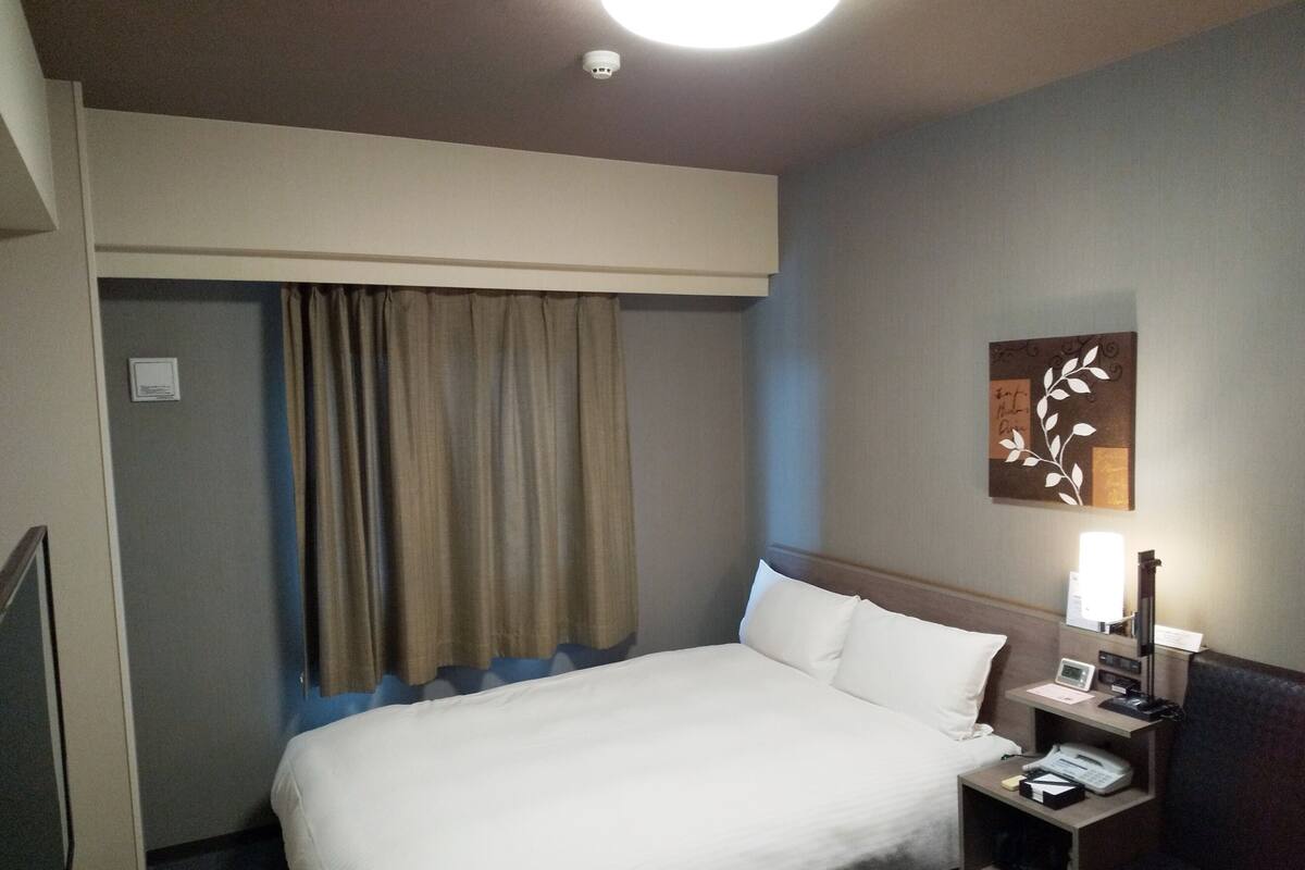Habitació d'hotel amb un llit, una tauleta de nit, una làmpada, una obra d'art penjada a la paret i una llum al sostre.