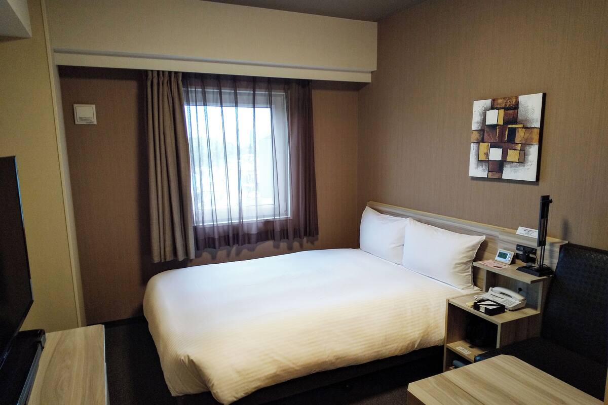 Habitació d'hotel amb un llit gran, una taula de nit amb un telèfon i una làmpada, un escriptori amb una cadira i una finestra amb cortines lleugeres.