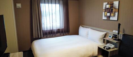 A hotel room with a large bed, a bedside table with a phone and a lamp, a desk with a chair, and a window with sheer curtains.