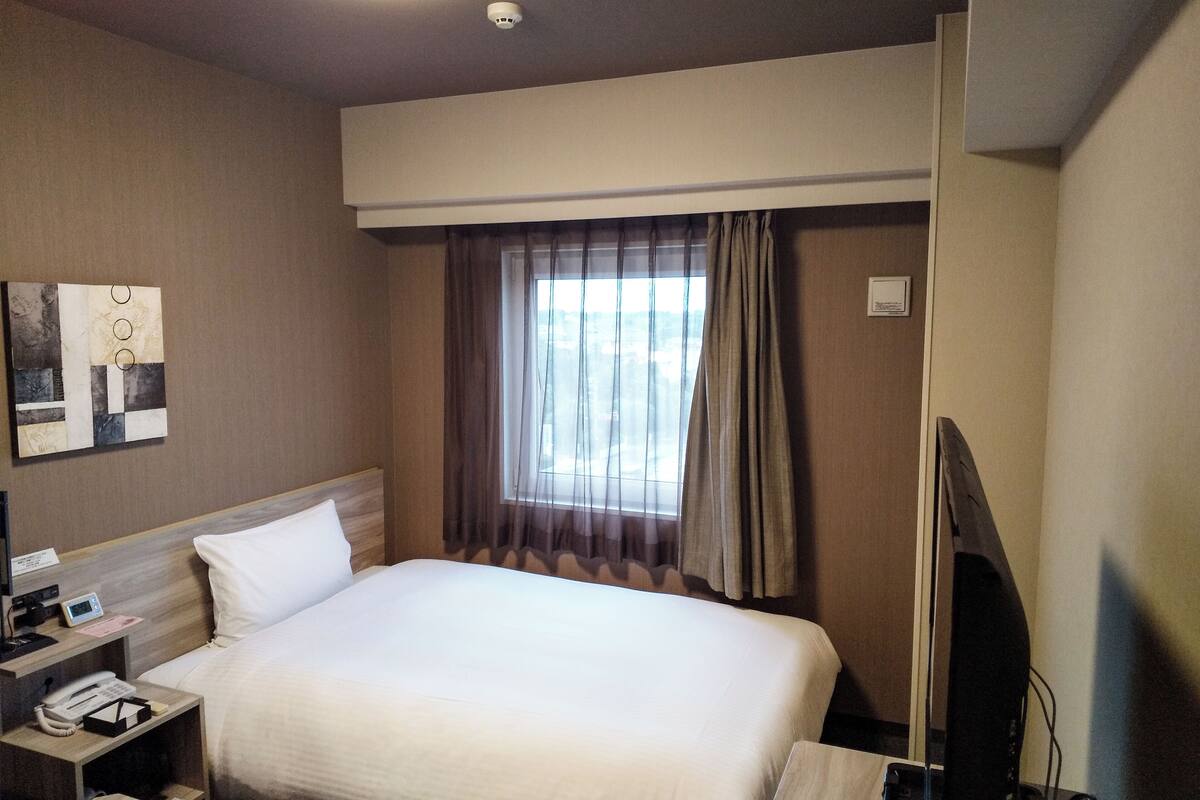 Habitació d'hotel amb llit, taula de nit, televisió i finestra amb cortines.