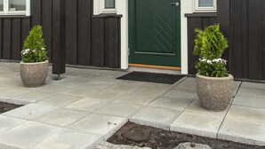 A dark wooden building with a green door, two potted plants, and a stone pathway leading to the entrance.