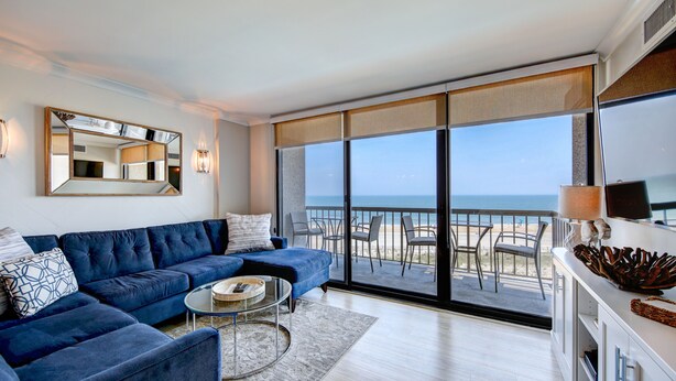 Un salon moderne avec un canapé bleu, une table basse ronde et une grande fenêtre donnant sur la plage.