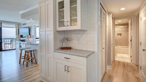 Une cuisine moderne avec des meubles blancs, un tabouret de bar en bois et un plan de travail agrémenté d’un vase décoratif.