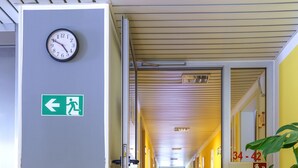 A hallway with a clock on the wall, an emergency exit sign, and a plant. - Motel Drei König (Heimsheim)