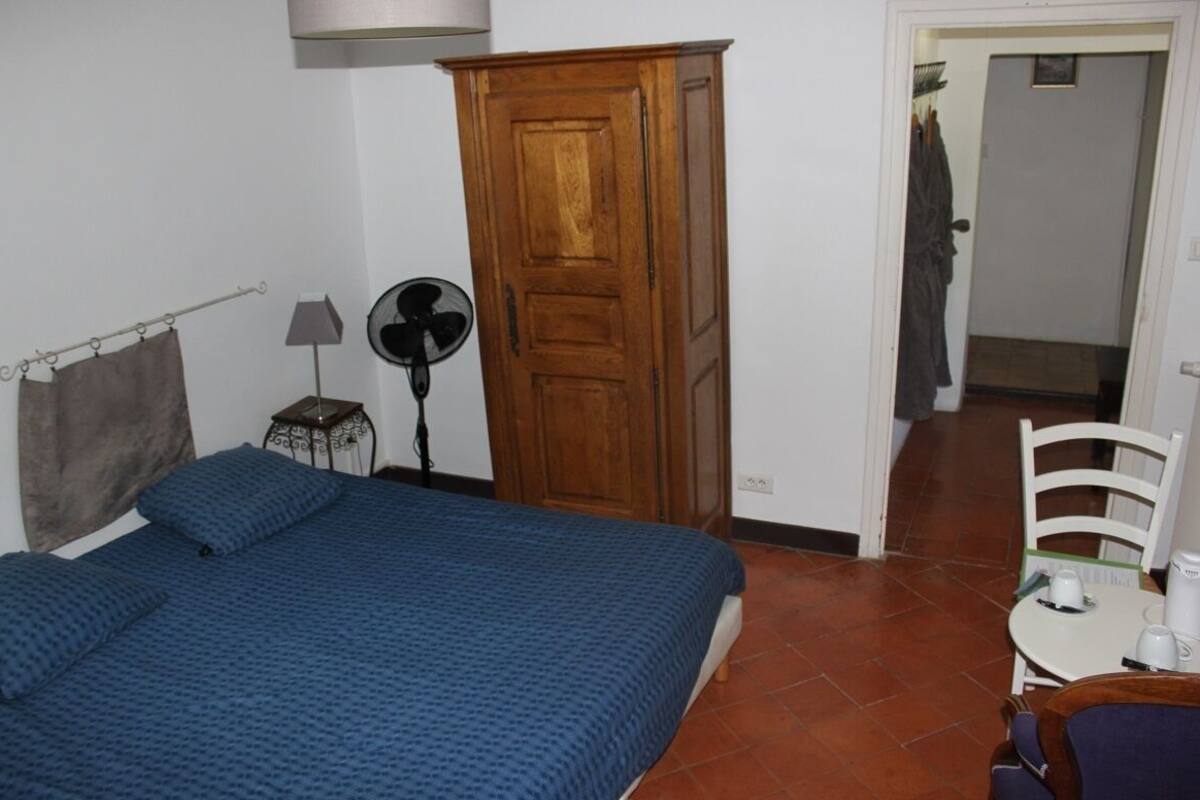 Een slaapkamer met een bed, een houten kast, een ventilator en een tafel met een stoel.