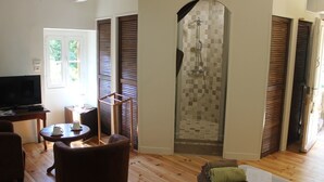 A living room with a television, a coffee table, and a sofa. There is a shower area with a tiled wall and wooden shutters. - Domaine de la Carrière (Sumene)