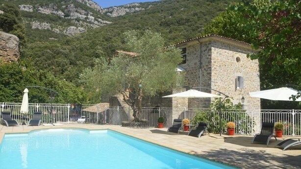 A pool area with lounge chairs and umbrellas, surrounded by stone buildings and a mountainous backdrop. - Domaine de la Carrière (Sumene)
