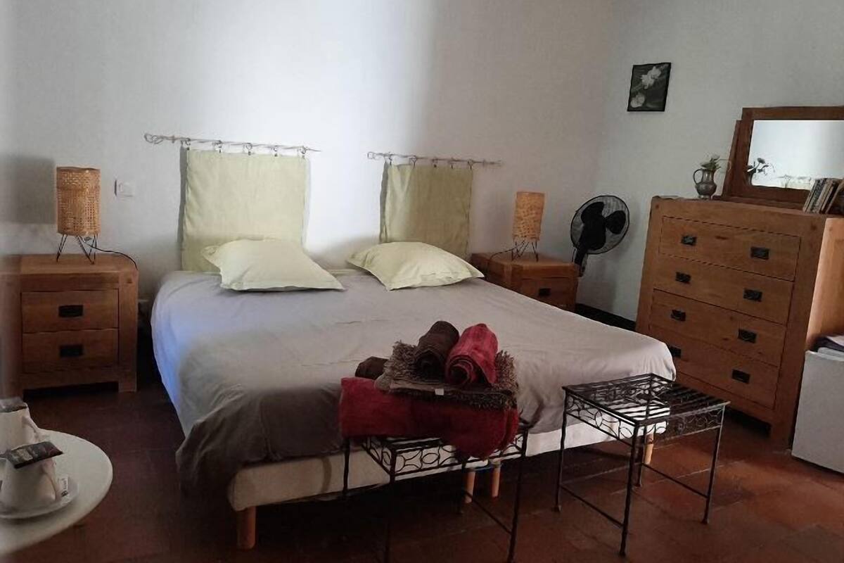 Een slaapkamer met een bed, twee nachtkastjes, een dressoir, een ventilator en een klein tafeltje met een stoel.