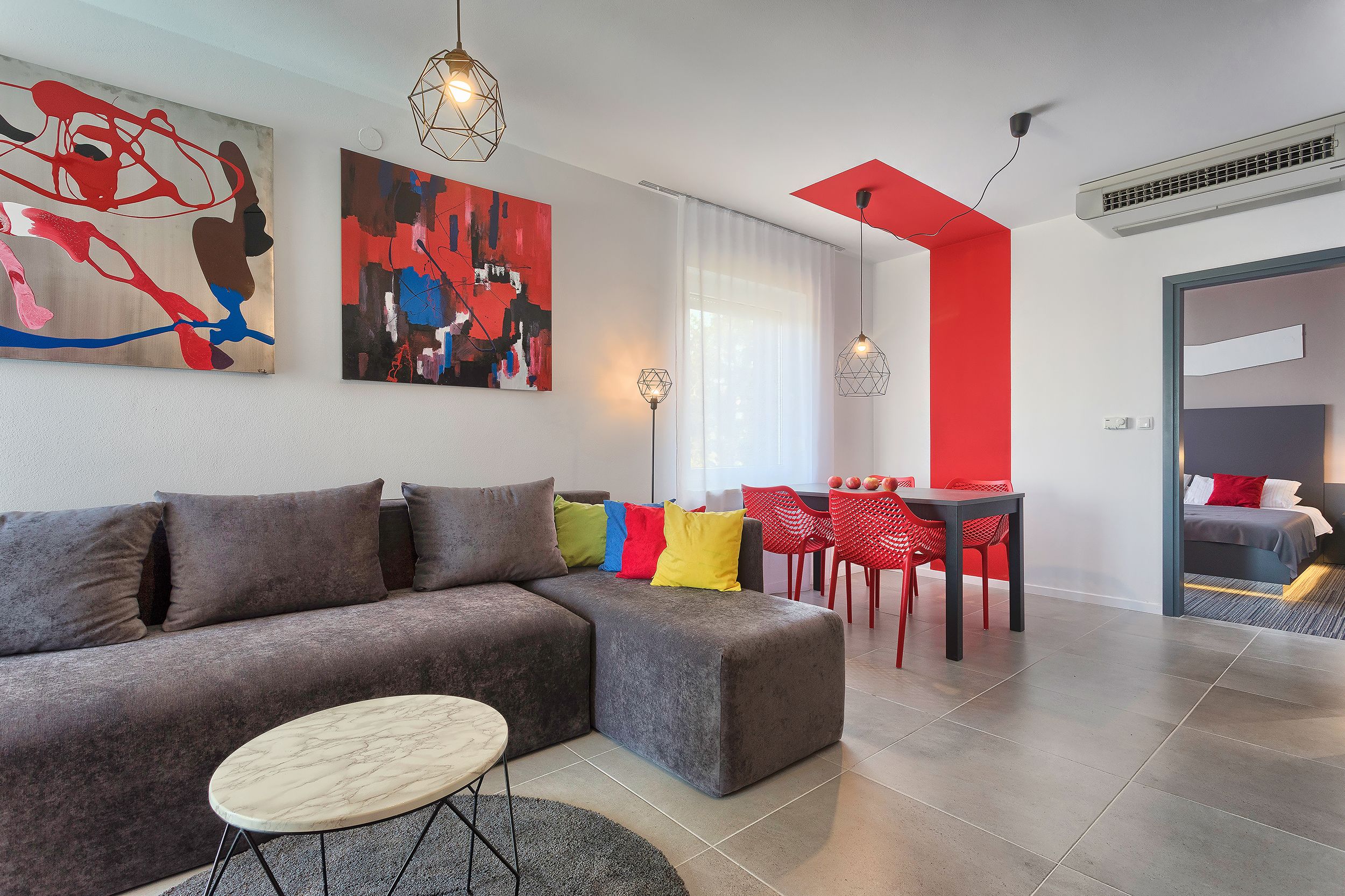 A modern living room with a grey sectional sofa, a round coffee table, and colorful cushions. There are abstract paintings on the walls, a dining area with red chairs, and a bed in the background.