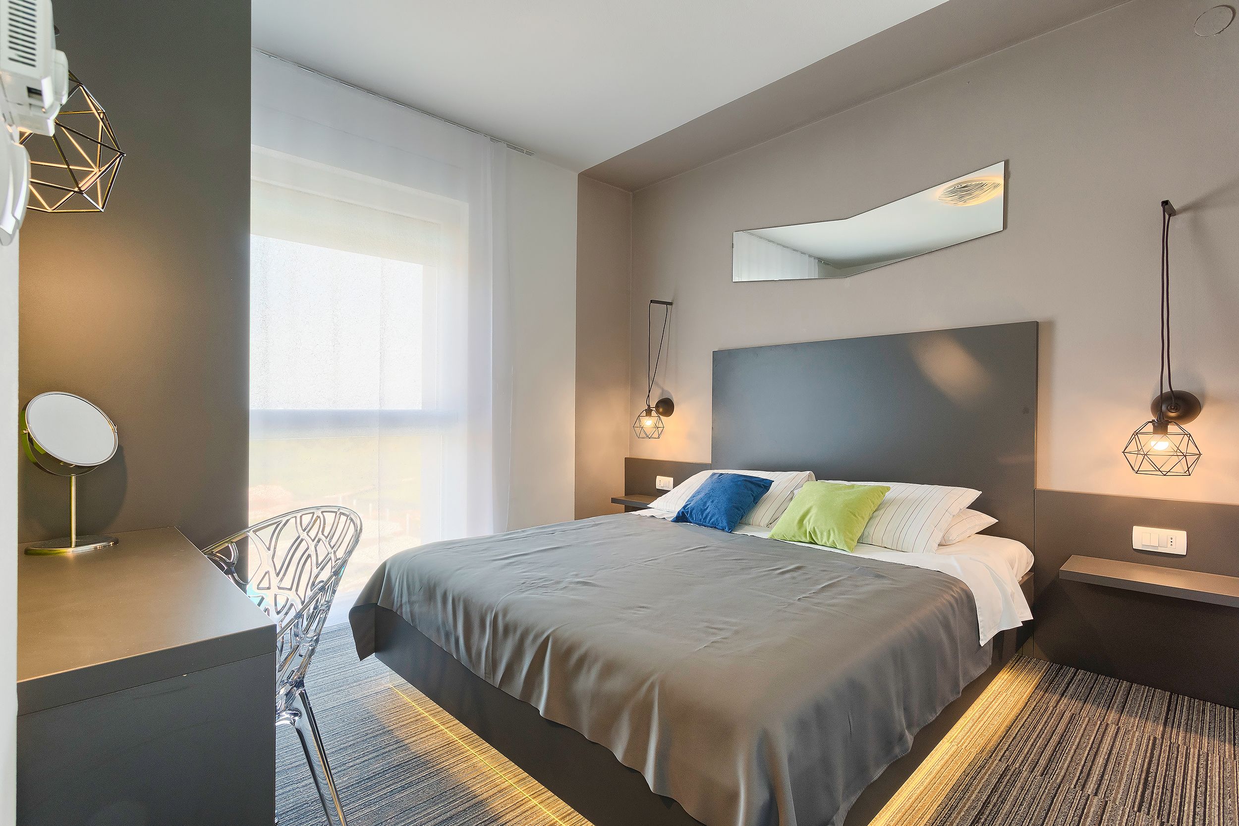 A modern hotel room with a large bed, a desk with a mirror, and a chair. There is a window providing natural light and a view of the city.