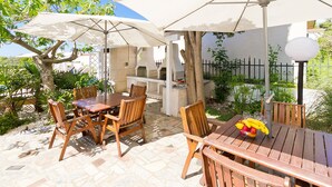 Une terrasse extérieure avec des tables et des chaises en bois, des parasols blancs et un espace barbecue.