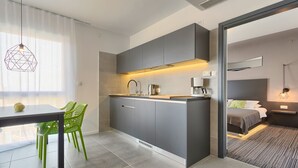 Une cuisine moderne avec des meubles gris, une table à manger entourée de chaises vertes, et une vue sur une chambre avec un lit et une table de chevet.