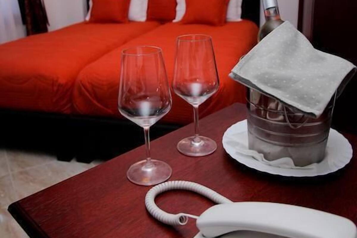 A hotel room with a bed, two wine glasses, a bottle of champagne, a telephone, and a painting on the wall.