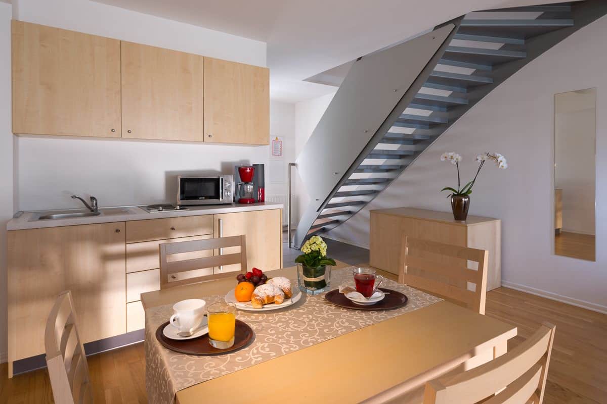 A modern kitchen with wooden cabinets, a microwave, and a coffee maker. There is a dining table with a vase of flowers and a tray with food and drinks.