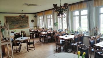 A dining area with wooden tables and chairs, large windows with curtains, and various potted plants.