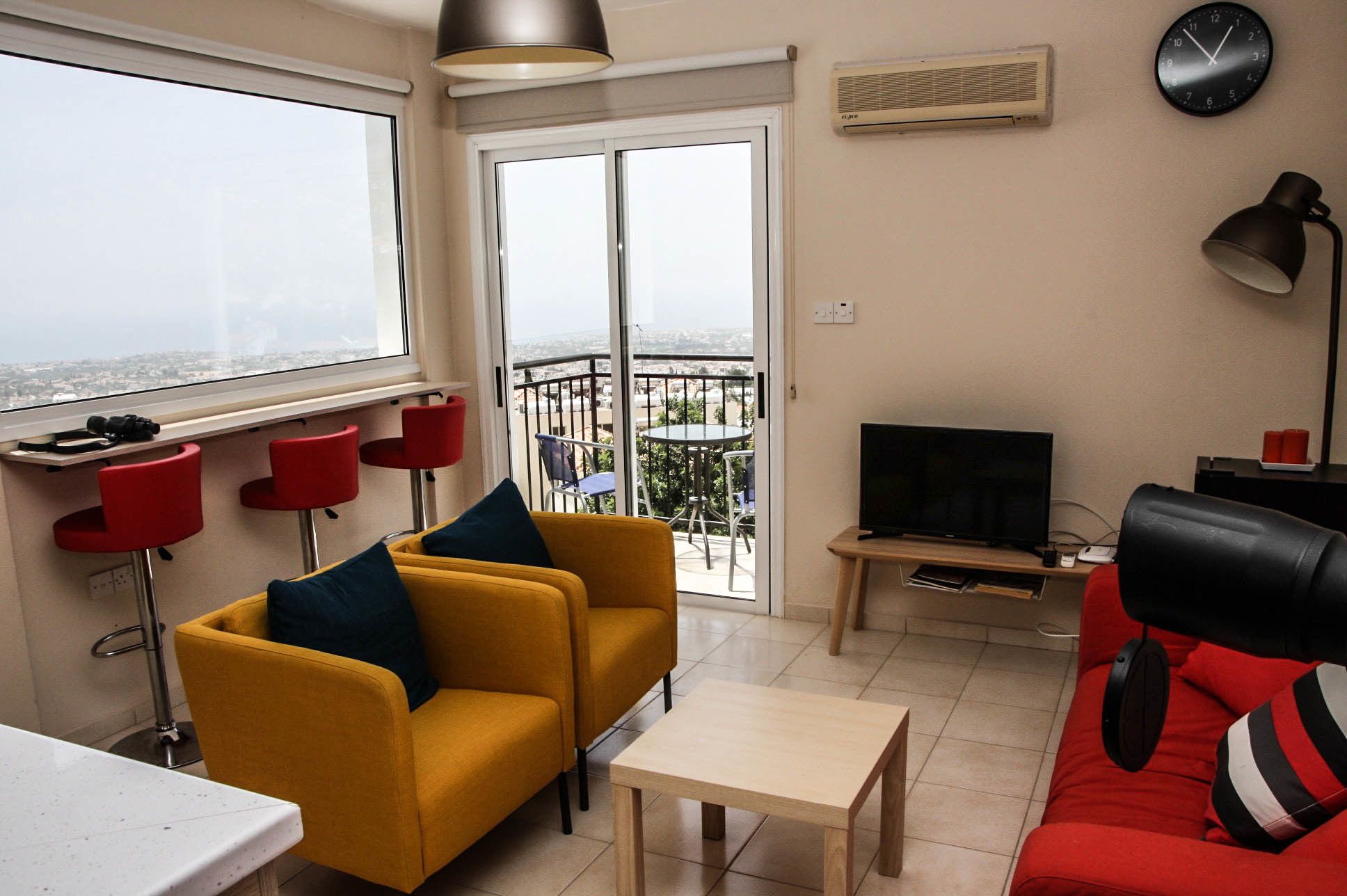 A modern living room with a yellow sofa, red chairs, a TV, and a balcony with a view.