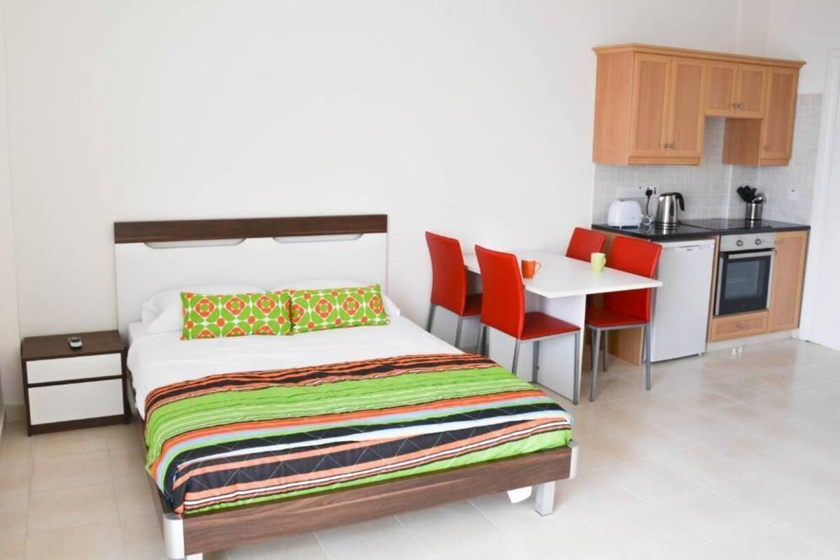 A bedroom with a bed, bedside table, and a small desk with a chair. There is a kitchenette with a stove, oven, and cabinets.
