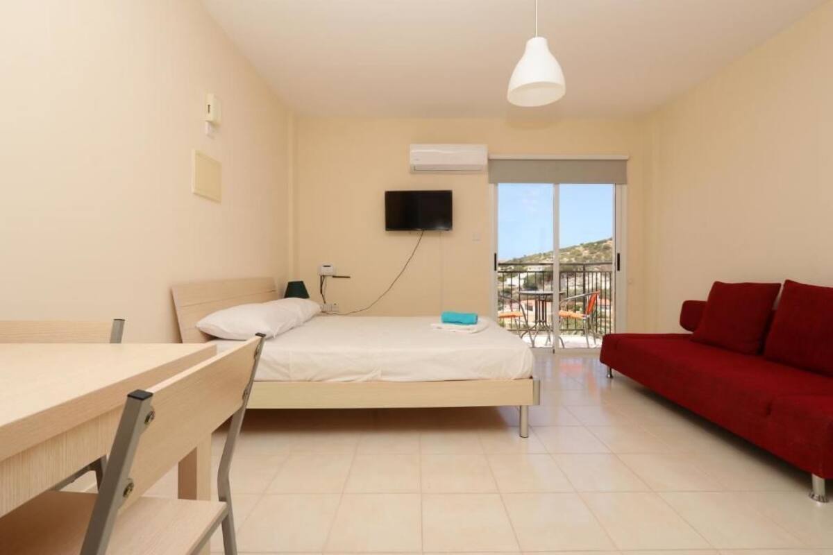 A bedroom with a bed, a desk, a red sofa, a TV, and a balcony with a view.