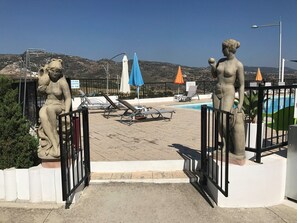 Zona de piscina con dos estatuas de piedra, sillas de descanso y sombrillas.