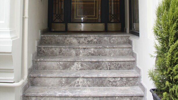 Una serie de escalones de mármol conducen a una puerta con marco oscuro y un panel de vidrio, una pequeña mesa con una lámpara y una planta en maceta al lado de las escaleras.