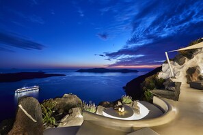 A sunset view from a seaside resort with a ship in the distance, a curved seating area, and rocky terrain. - Villa Etheras (Santorini)
