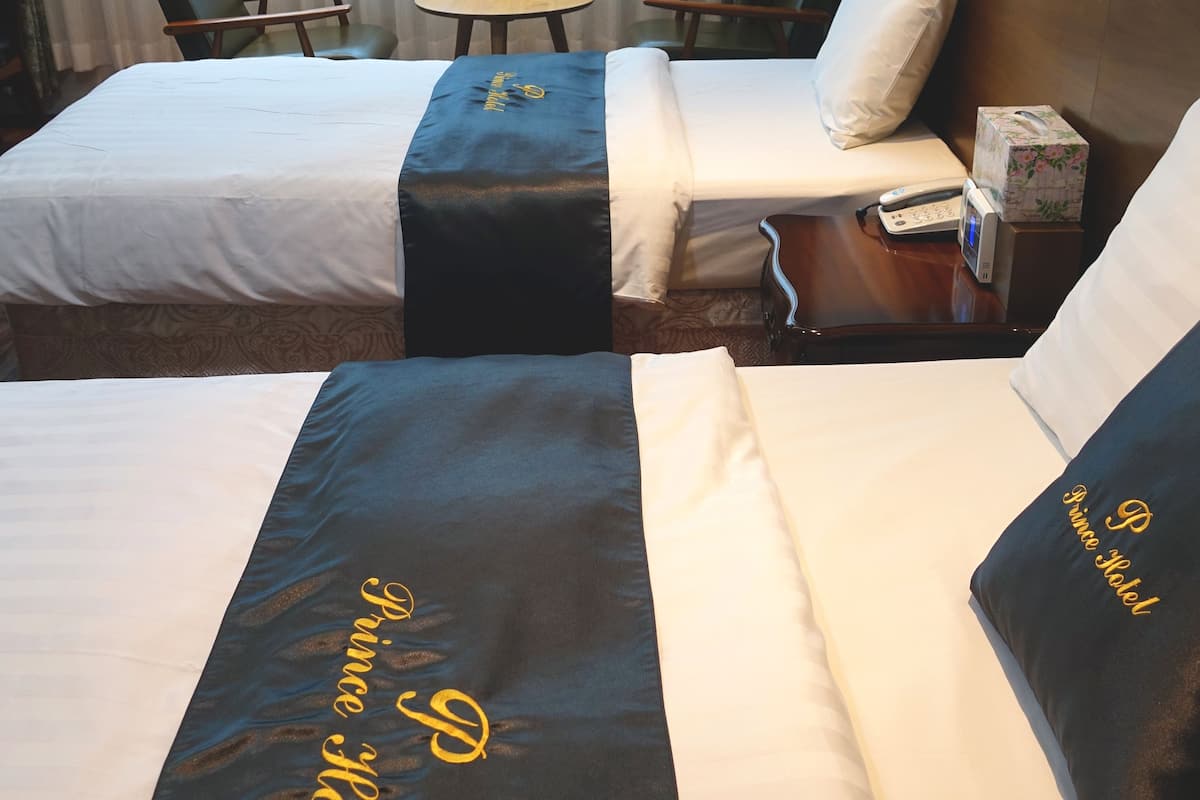 A hotel room with two beds, a small table, a chair, and a bedside table with a phone and a box.