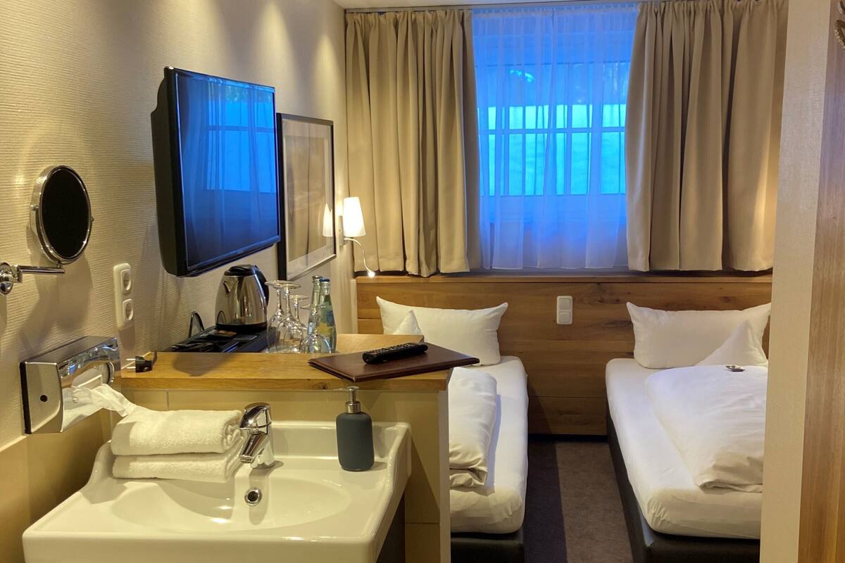 A hotel room with a large bed, a smaller bed, a vanity with a sink, a television, and a window with curtains.