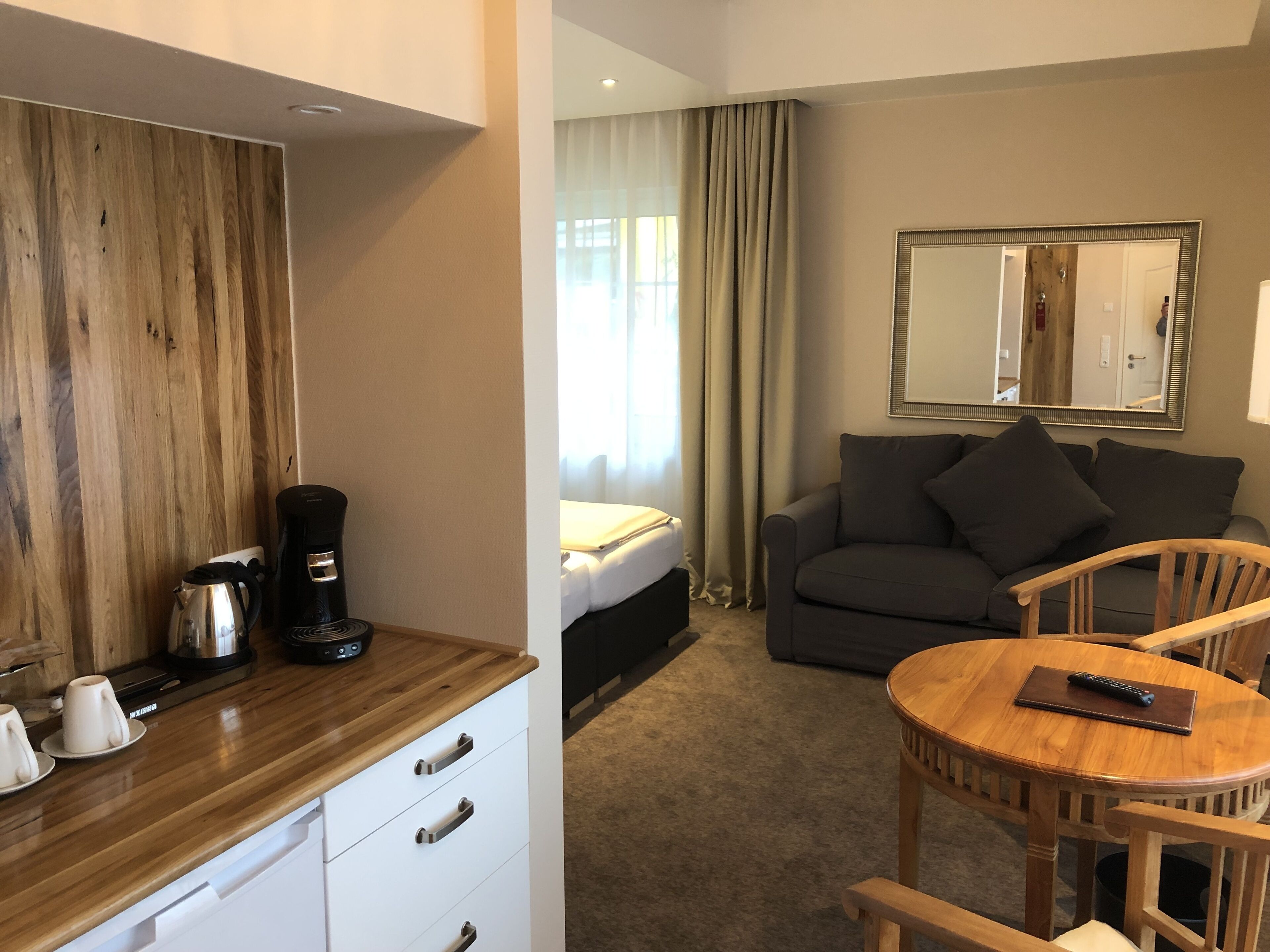 Ein Hotelzimmer mit einem Holztisch, einer Kaffeemaschine, einem Sofa, einem runden Holztisch und einem Spiegel an der Wand.