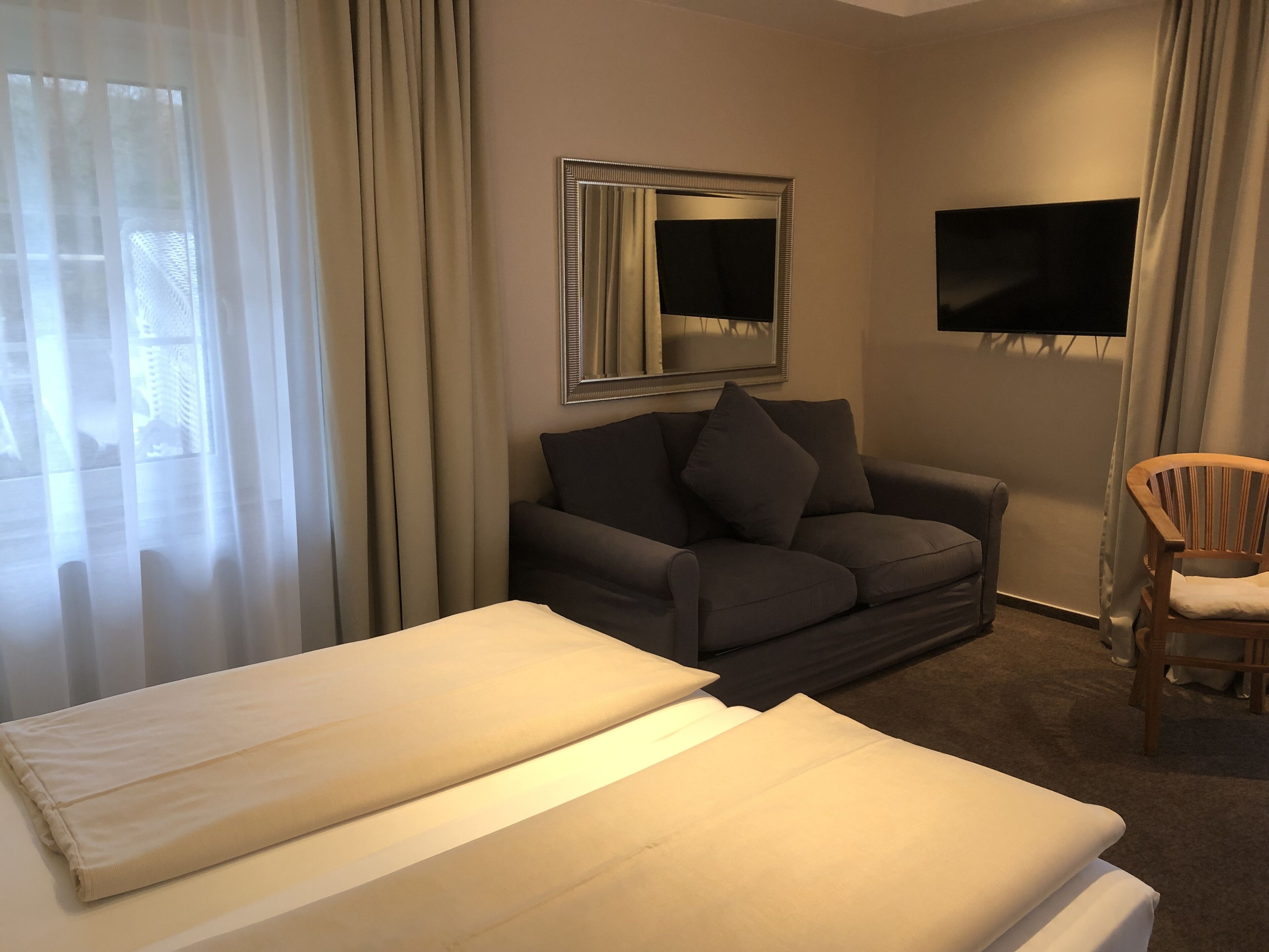 A hotel room with a bed, a sofa, a wooden chair, a TV mounted on the wall, and a window with curtains.