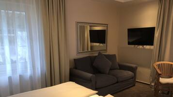 Ein Hotelzimmer mit Bett, einer Couch, einem Holzsessel, einem an der Wand montierten Fernseher und einem Fenster mit Vorhängen.