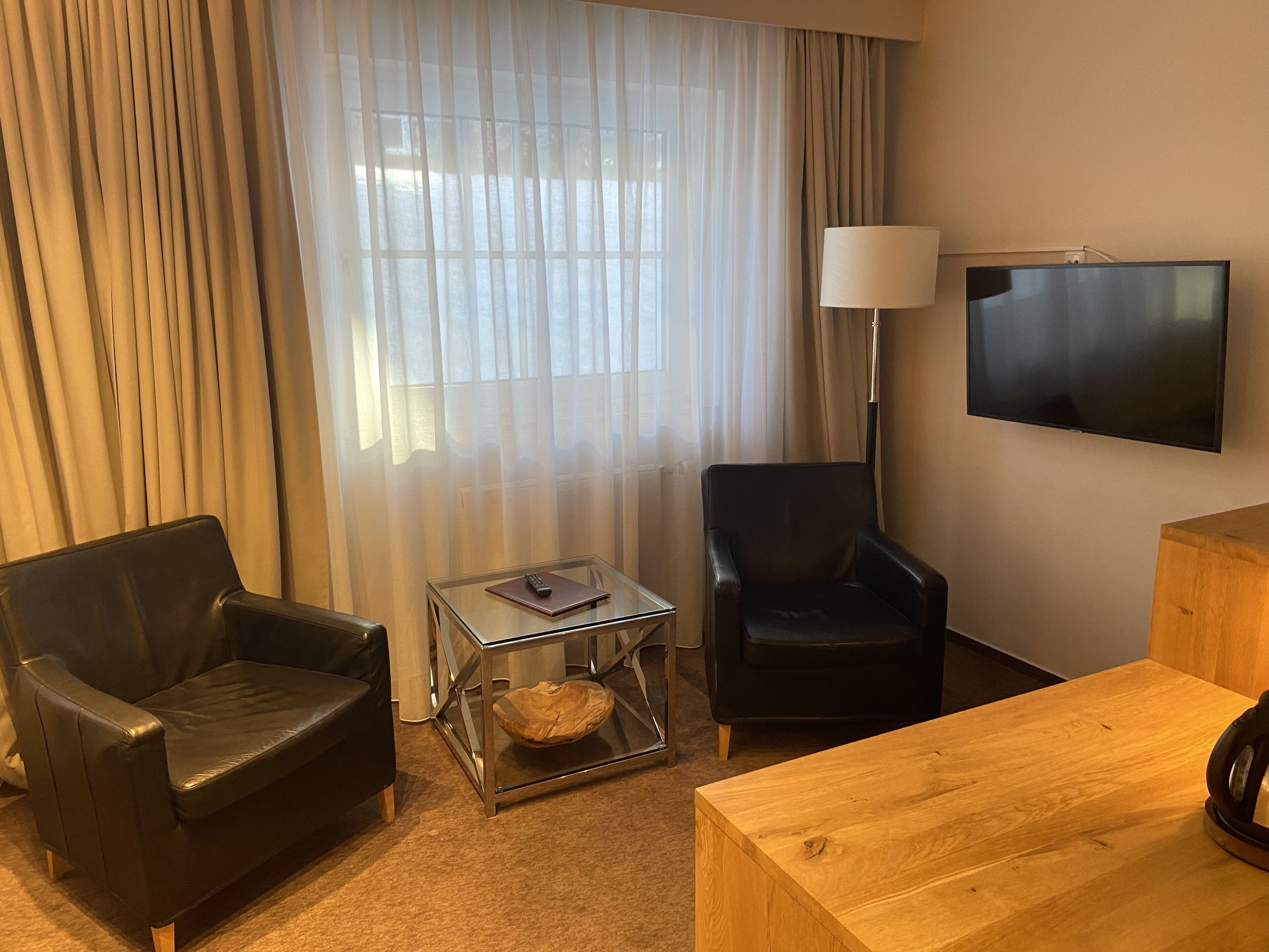 Una camera d'hotel con un divano in pelle nera, un tavolino in legno, un tavolino di vetro con un telecomando, una poltrona nera, una lampada e una TV a parete.