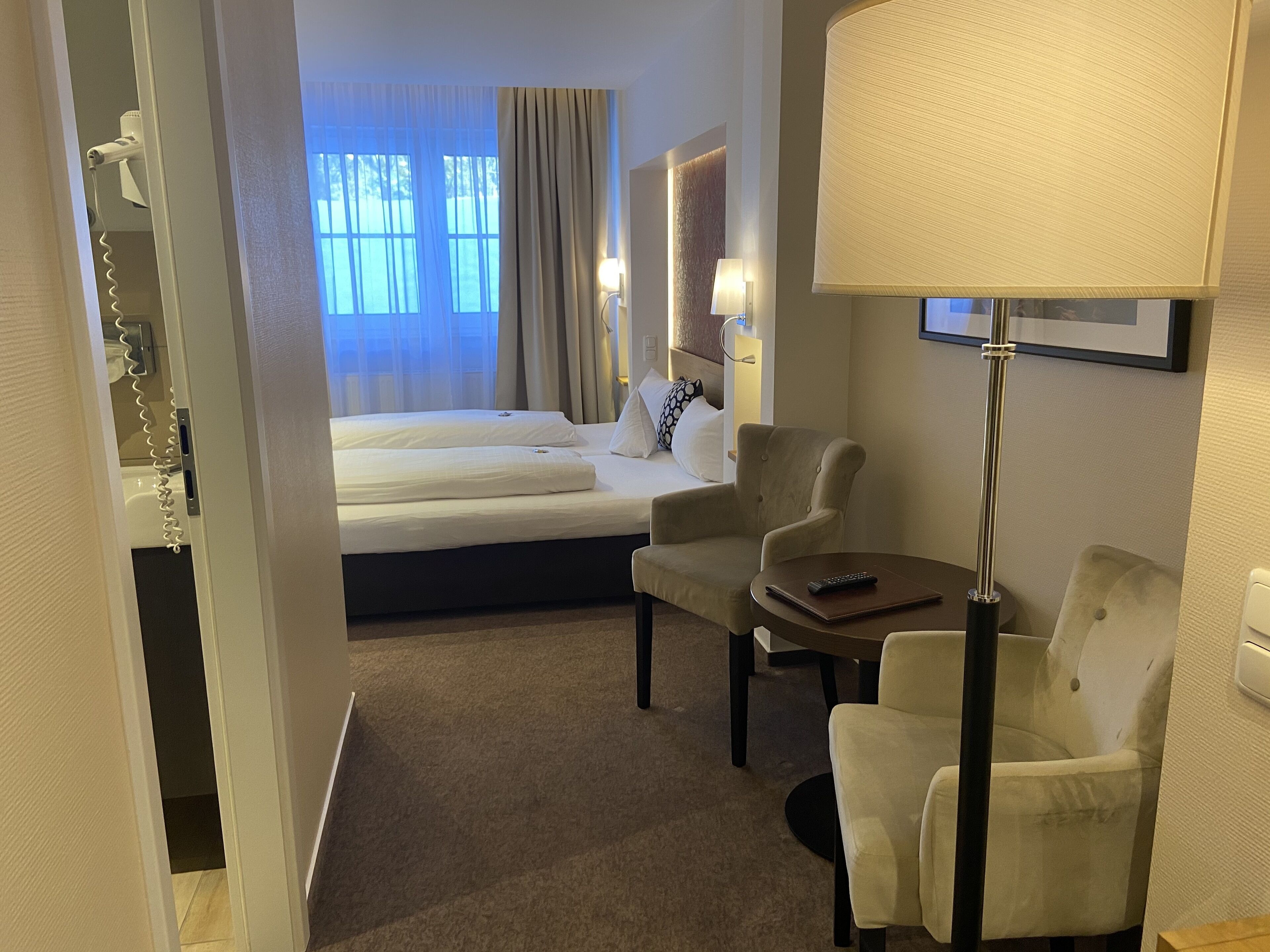 Une chambre d’hôtel avec un lit, deux fauteuils, une petite table, une lampe et une fenêtre avec des rideaux.