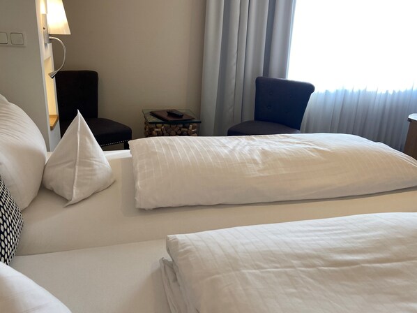 A hotel room with a bed, pillows, a chair, a small table with a remote control, and a lamp.