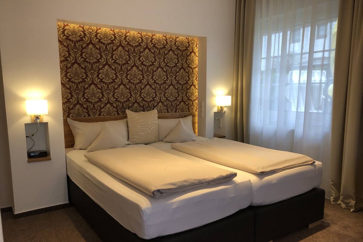 A double bed with a patterned headboard, two bedside lamps, and a window with curtains.