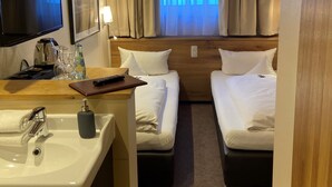 A hotel room with two beds, a desk with a sink, a television, and a window with curtains. - Hotel Petersen‘s Landhaus Scharbeutz (Scharbeutz)