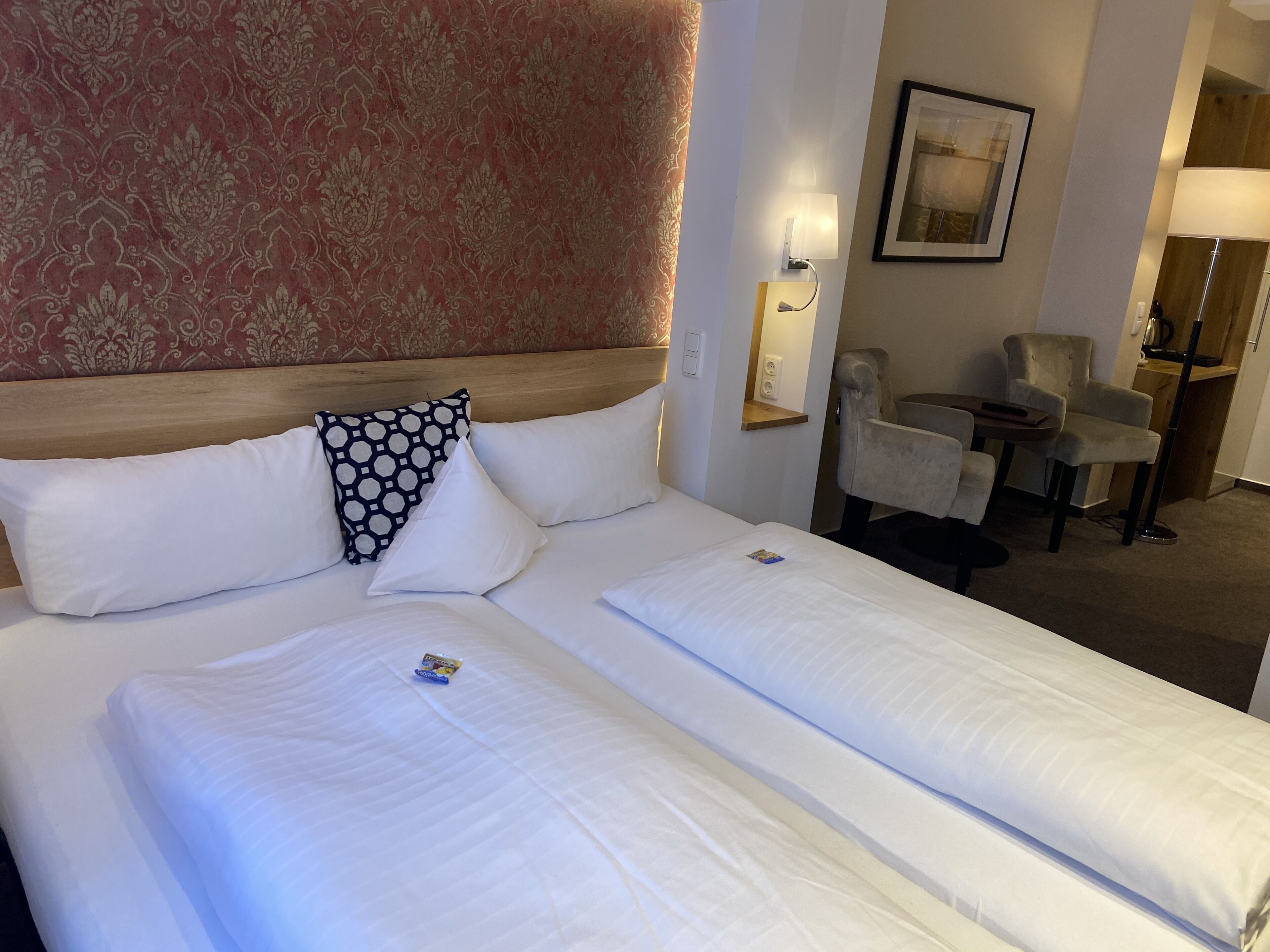 A hotel room with a large bed, two pillows, a headboard, a wall-mounted lamp, a framed picture, and a seating area with a chair and a small table.