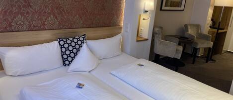 A hotel room with a large bed, two pillows, a headboard, a wall-mounted lamp, a framed picture, and a seating area with a chair and a small table.