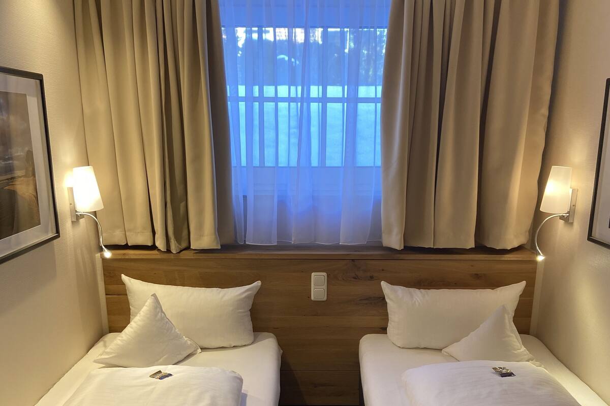 A hotel room with two beds, a window with curtains, wall-mounted lights, and a framed picture on the wall.