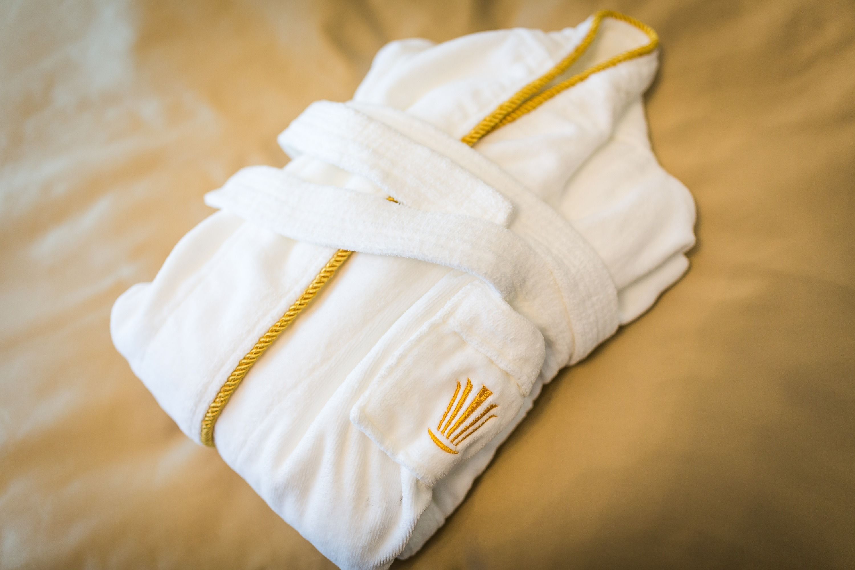 Een witte handdoek met gouden bies en een gevlochten gouden handgreep, netjes gevouwen op een bed.