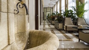 Um banco de pedra com encosto curvo, uma parede de pedra com um acessório de metal e um corredor com cadeiras e mesas, sugerindo uma área de lounge.