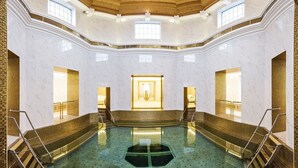 Uma grande piscina interna com paredes de mármore e detalhes dourados, cercada por degraus e corrimãos.