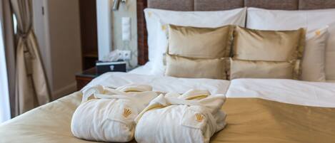 A neatly made bed with white linens and two towels with a gold emblem, a wooden headboard, and a wall-mounted lamp.