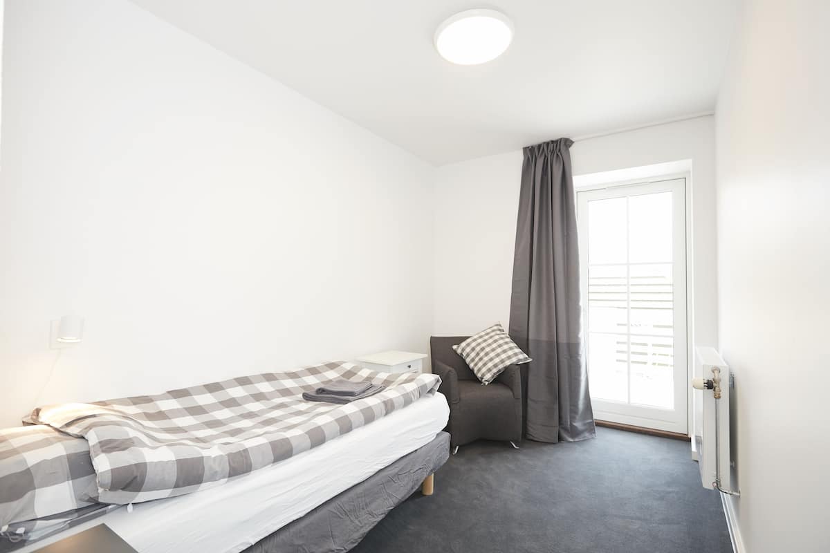 A small, single-bed room with a grey checkered bedspread, a grey armchair with a checkered pillow, a window with grey curtains, and a white radiator.