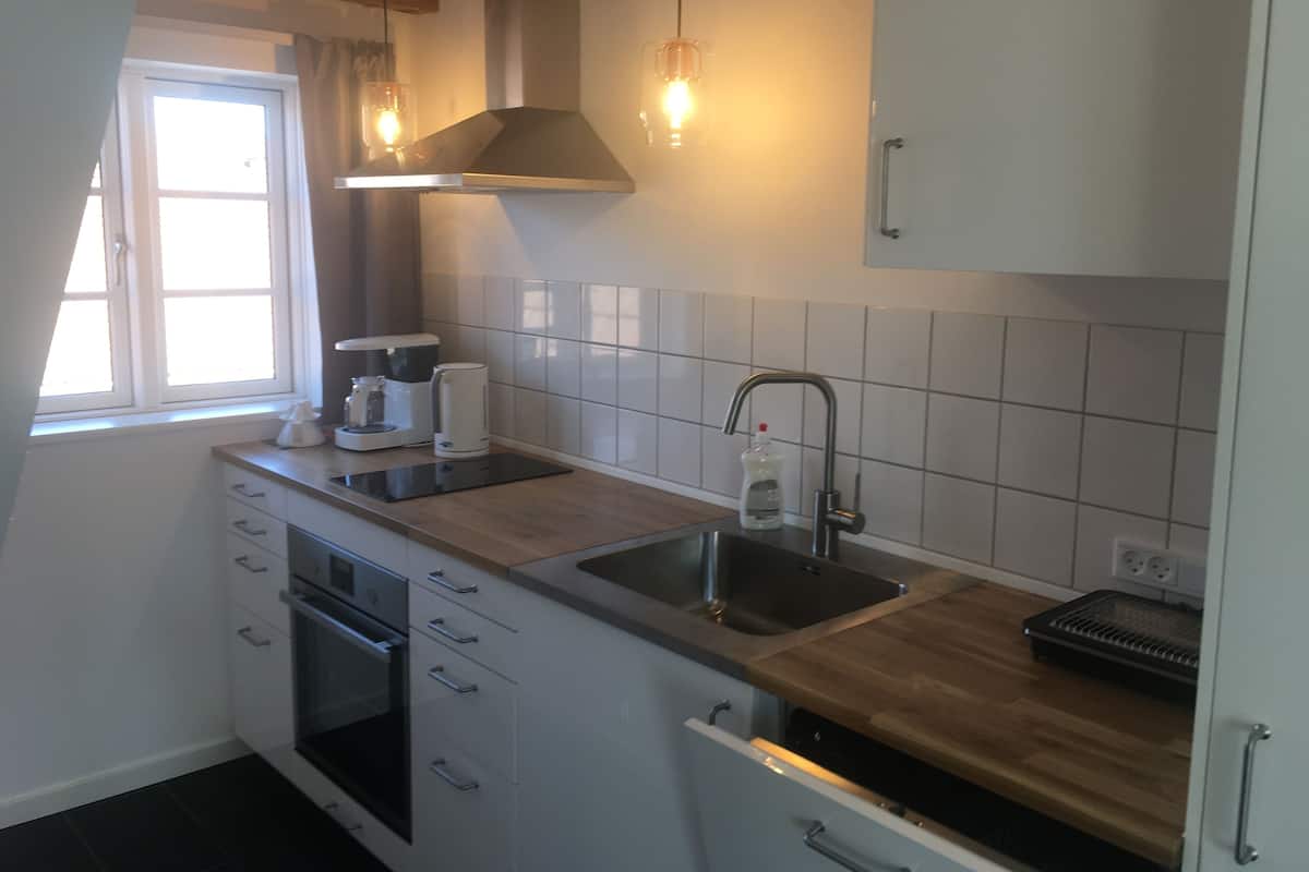 A modern kitchen with white cabinets, a double sink, and a built-in oven. There is a window providing natural light and a hanging light fixture above the sink.