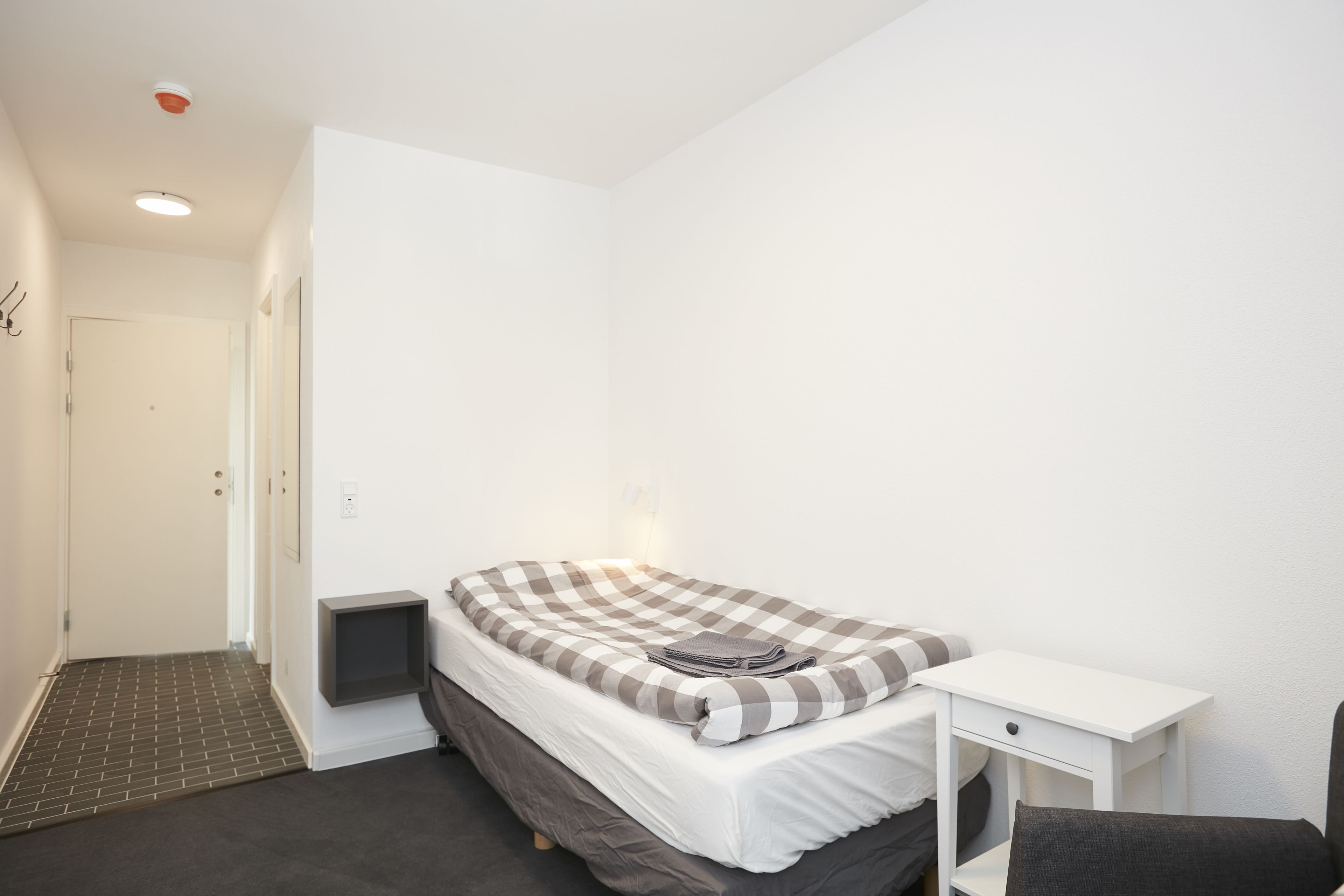 Een kleine, schone kamer met een eenpersoonsbed, een nachtkastje en een stoel.