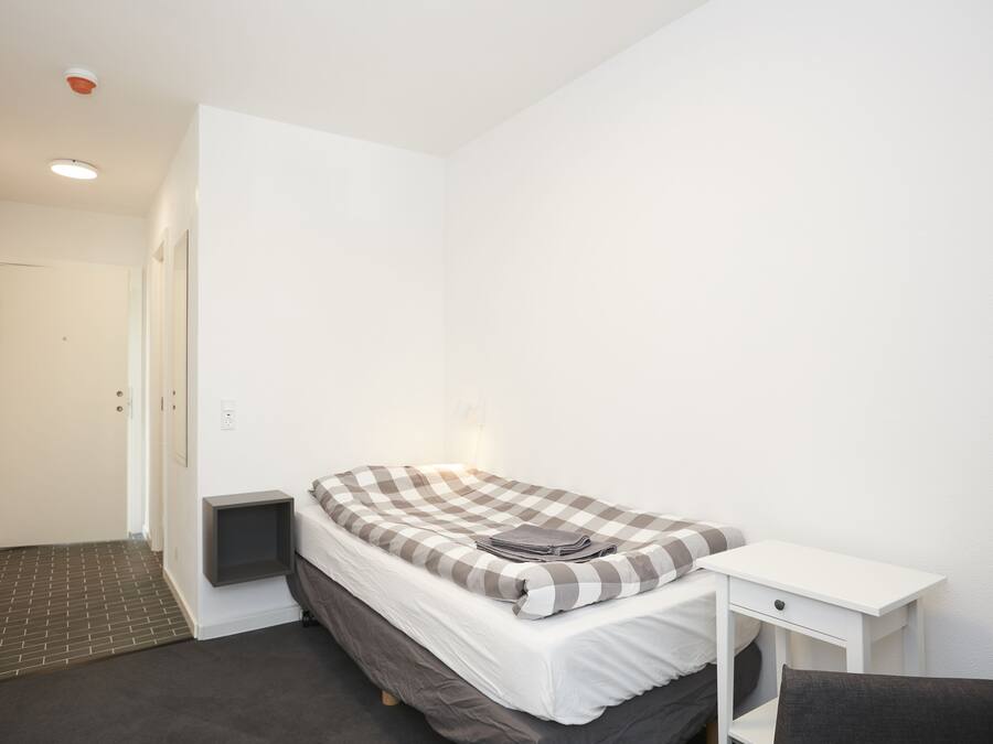 Een kleine, schone kamer met een eenpersoonsbed, een nachtkastje en een stoel.