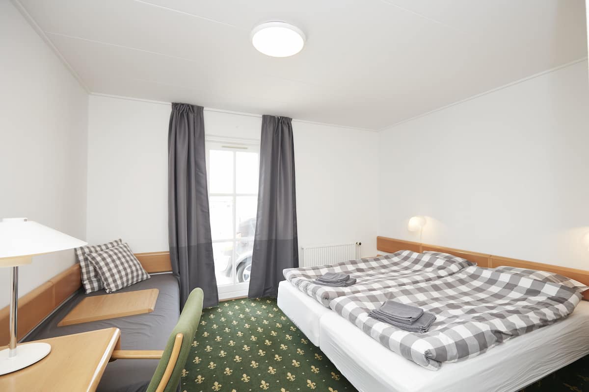 A hotel room with a bed, a desk, a chair, a window with curtains, and a radiator.