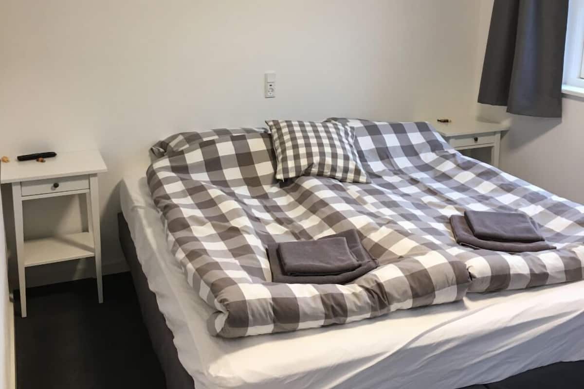 A single bed with a checkered bedspread, two pillows, and two folded towels. There is a white nightstand to the left and a white dresser to the right.