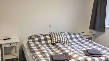 A single bed with a checkered bedspread, two pillows, and two folded towels. There is a white nightstand to the left and a white dresser to the right.